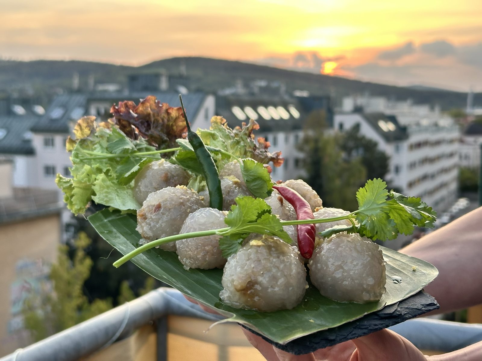 Gefüllte Tapioka-Klößchen auf Bananenblatt mit Salat, Koriander und Chili, im Hintergrund Wiener Dächer bei Sonnenuntergang